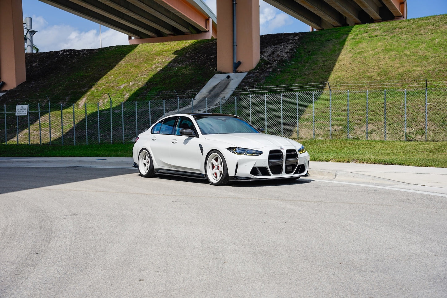White BMW car on a road with a grassy hill and overpass in the background